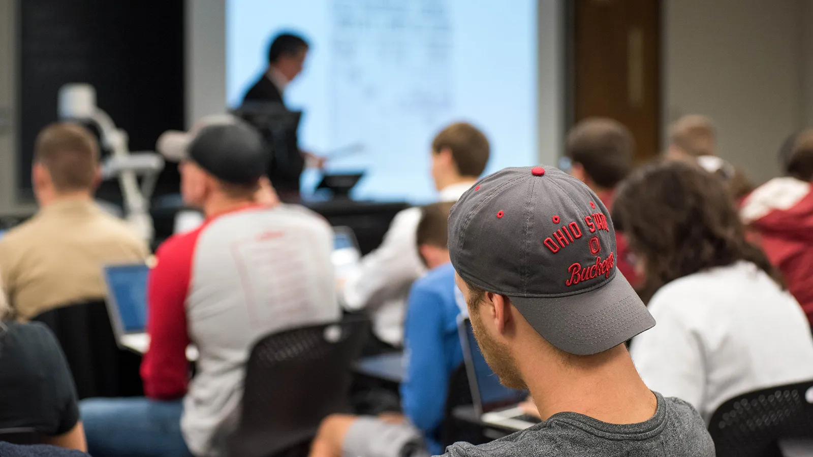 Student sitting in the back of class with an Ohio State ballcap takes notes as Bruno Cabanes lectures